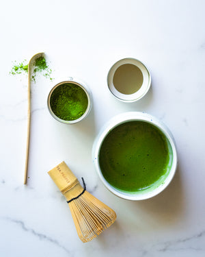 Matcha green tea in a bowl with a matcha whisk and an opened tin of matcha